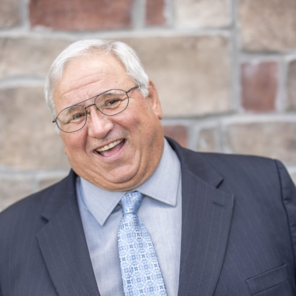 Rick Sackman, Regional Plant Operations Coordinator at Arrow Senior Living Home Office, smiling in a professional headshot with short white hair, glasses, a dark suit, and a blue patterned tie against a stone wall background.