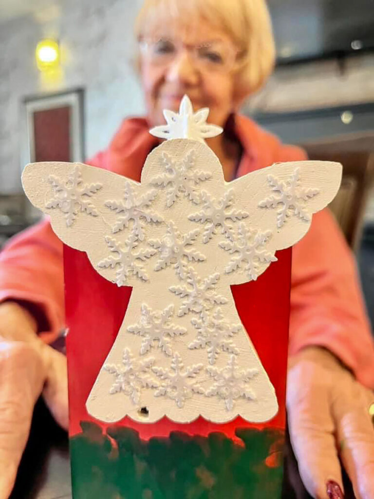 A smiling senior woman holds a decorated art craft of angle with snowflakes on top.