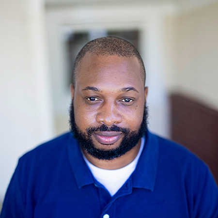 Aaron Blevins, Regional Wellness Coordinator for Arrow Senior Living, wearing a navy blue polo shirt and smiling slightly while standing in a bright hallway.