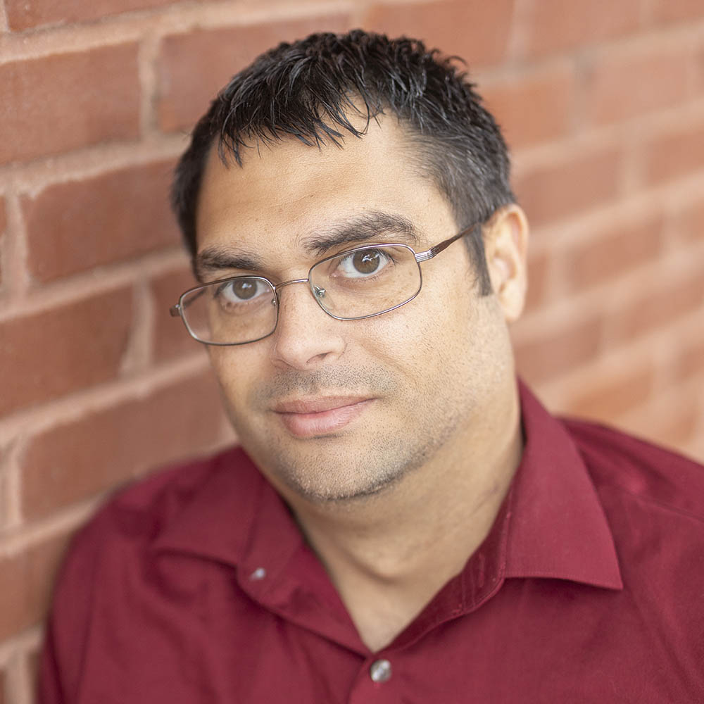 Ariel Forste of Arrow Senior Living, wearing a red shirt and glasses, leaning against a brick wall while looking calmly at the camera with a slight smile.