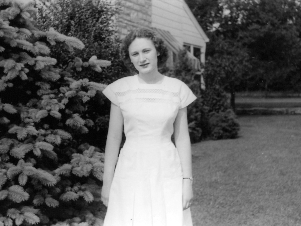 In 1951 a past photo of Joan, she has almost shoulder-length wavy hair and is standing outside by a house in a short-sleeved dress with lace.