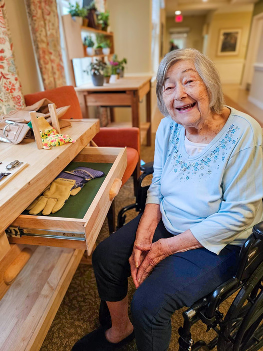 Elderly woman in a light blue shirt sitting in a wheelchair, smiling and opening a drawer filled with gardening tools in a well-lit room.