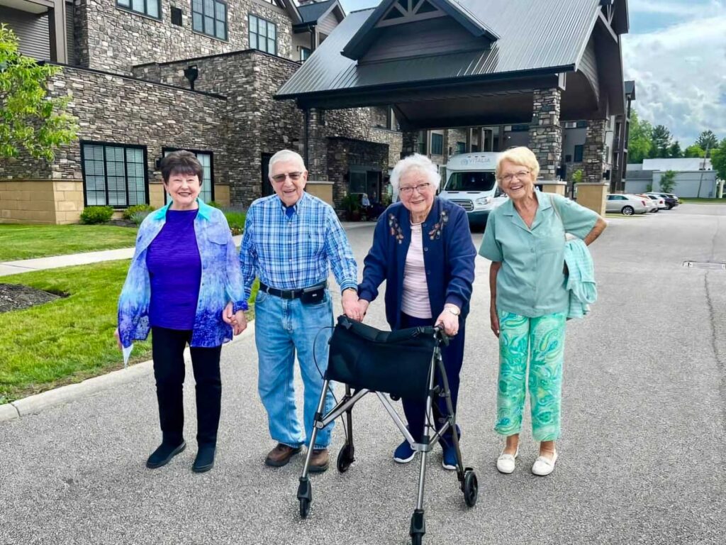 Four senior residents enjoying a walk outside a senior community building, smiling and holding hands, with one using a walker.