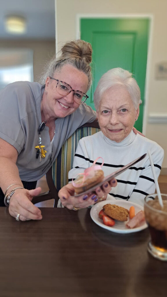 Senior resident and staff member smiling together at a community event, with the resident holding a small pastry wrapped with a ribbon, enjoying snacks and a drink at the table.