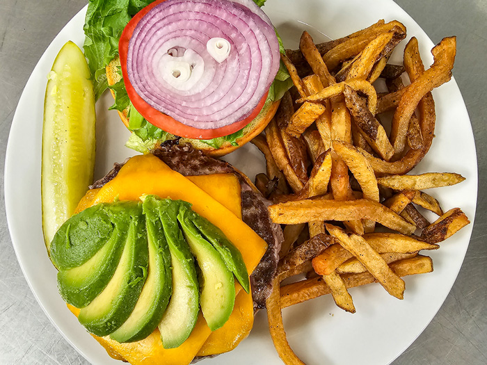 Delicious avocado cheeseburger topped with cheddar cheese, lettuce, tomato, and red onion, served with a side of crispy fries and a pickle spear on a white plate.