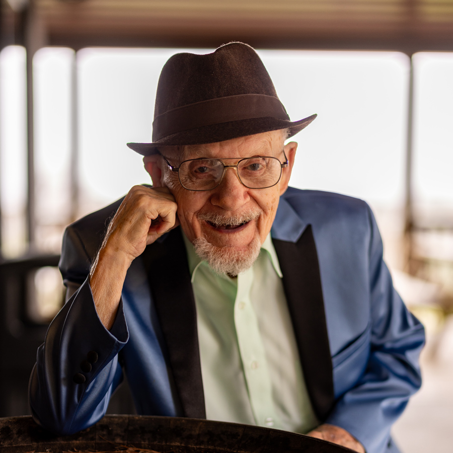 Smiling senior resident dressed in a stylish blue blazer and hat, leaning on a barrel during an outdoor event at a senior living community, capturing a moment of joy and charm.