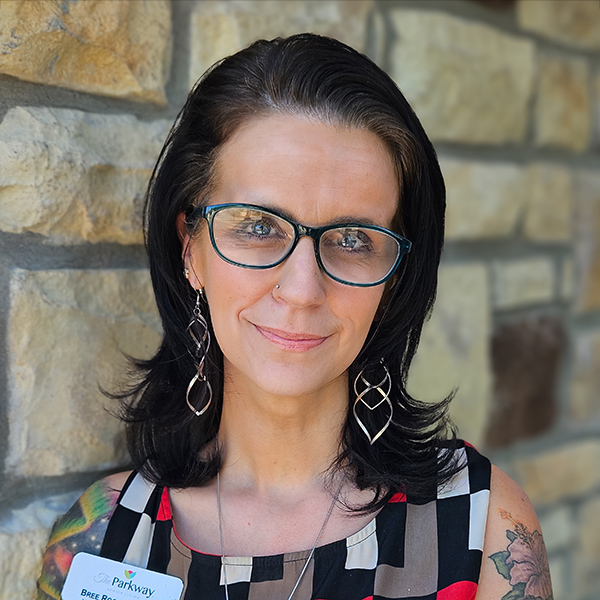Bree Rogers, Arrow Regional Memory Care Coordinator, poses outdoors against a stone wall, wearing glasses, statement earrings, and a patterned dress.