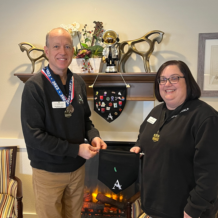 Two senior living employees stand in front of a decorated fireplace, holding an Employee Appreciation pin banner featuring various enamel pins and awards.