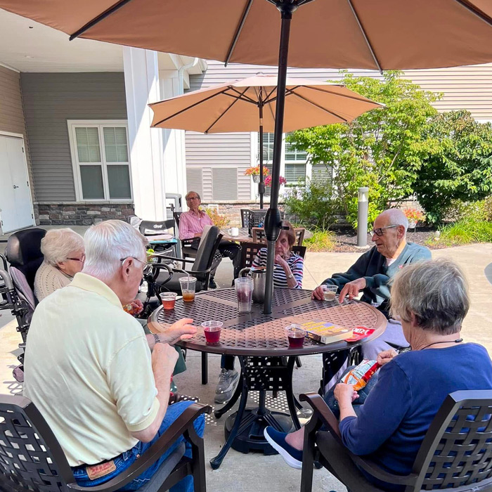 Senior residents sit under patio umbrellas enjoying drinks, snacks, and a game of bingo during a sunny spring afternoon in the community courtyard.