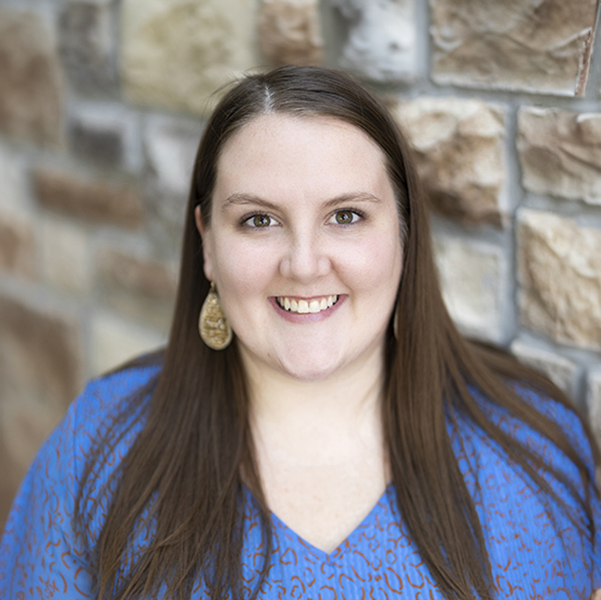 Betsy Baker, Regional Sales Coordinator at Arrow Senior Living, smiles warmly in a blue patterned blouse while standing in front of a neutral stone wall backdrop.