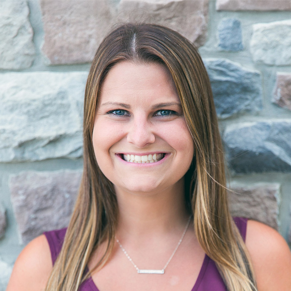 Bailey Brasdovich, Regional Director of Sales at Arrow Senior Living, smiles in a sleeveless plum top and necklace, standing before a multicolored stone wall.