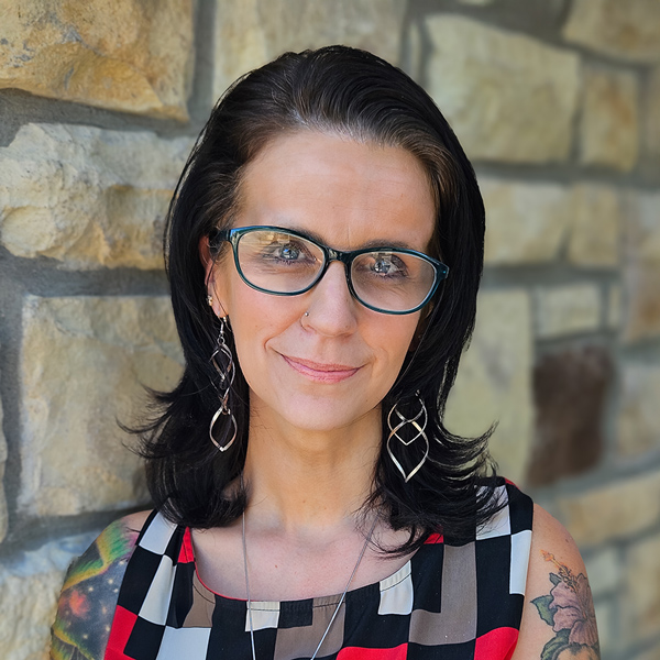 Bree Rogers, Regional Memory Care Coordinator at Arrow Senior Living, wearing glasses and patterned attire, standing by a stone wall.