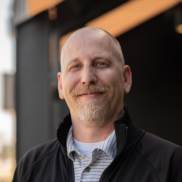 Brian Albrecht, Regional Culinary Director at Arrow Senior Living, smiling outdoors in a zip-up jacket and striped shirt with a blurred background of dark architecture.