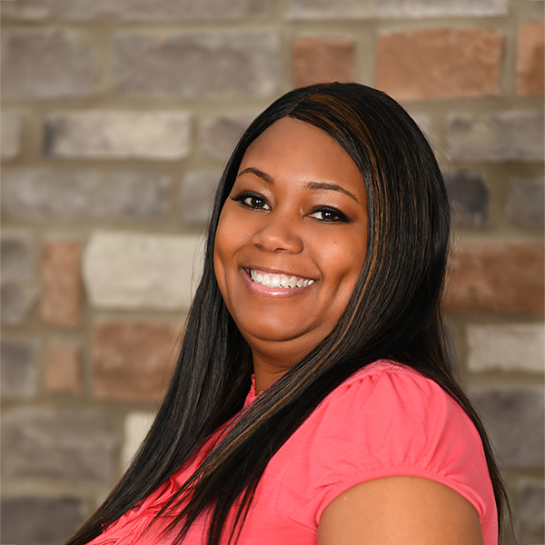 Che'Rri Colbert, Regional Business Office Director at Arrow Senior Living, smiling in front of a stone wall while wearing a coral blouse.
