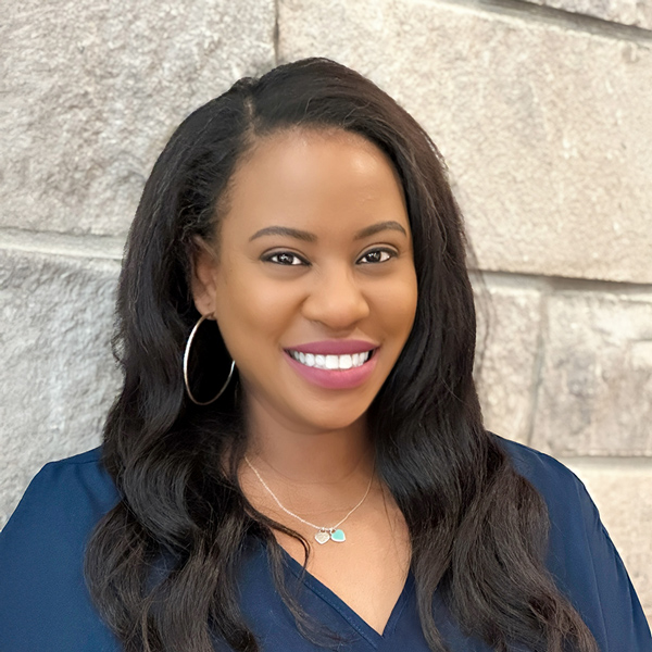 Danyelle Little, Resident Experience Coordinator at Arrow Senior Living, smiles warmly in a navy blouse with hoop earrings against a light stone wall backdrop.