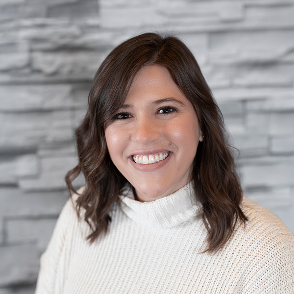 Paisli DiRuzza, Managing Director of Sales at Arrow Senior Living, smiles in a white turtleneck sweater, posed against a soft gray stacked stone wall backdrop.
