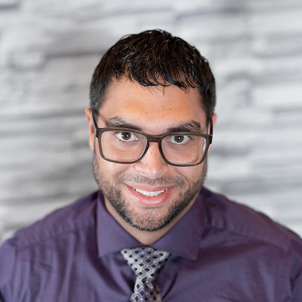 Ariel Forste, Senior Managing Director of Internal Operations at Arrow Senior Living, smiling in a purple shirt and tie in front of a light stone wall.