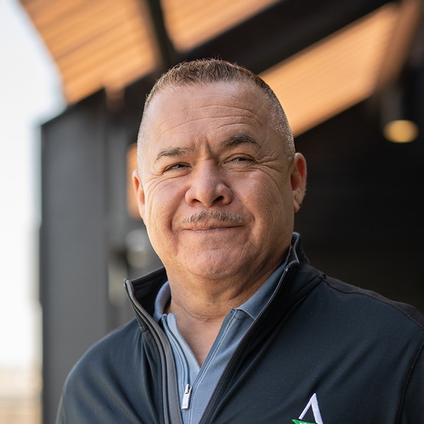 Humberto Barraza, Regional Director of Culinary Compliance at Arrow Senior Living, smiles while standing outdoors in a branded jacket with blurred architecture behind.