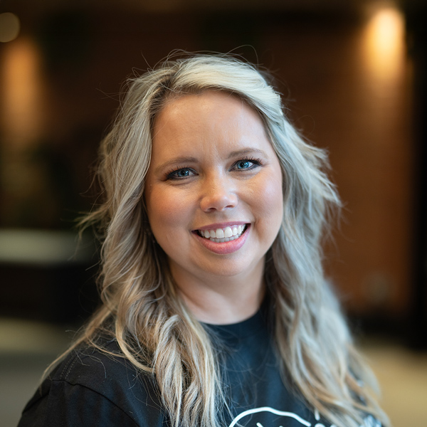 Kayla Barlow, Vice President of Resident Experience at Arrow Senior Living, smiles in a casual black shirt with wavy blonde hair in a warmly lit indoor setting.