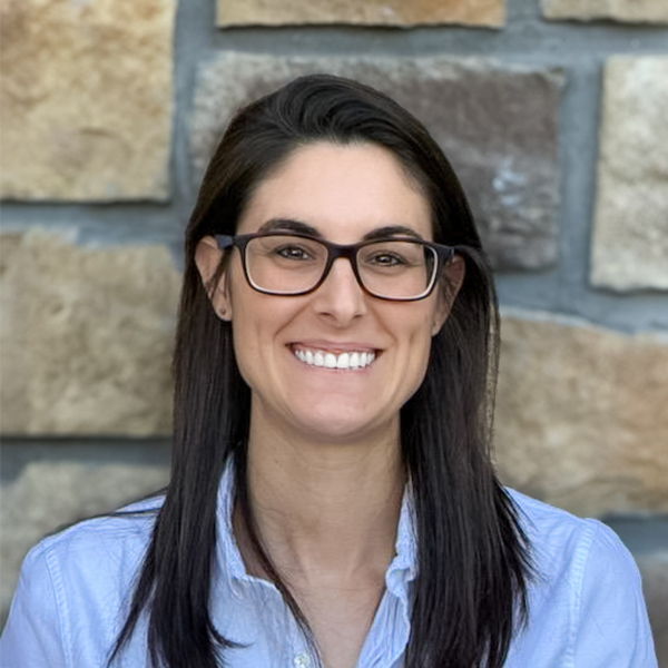 Kelsee Klein, Engagement Specialist at Arrow Senior Living, smiles while wearing glasses and a light blue button-up shirt, standing in front of a stone wall.