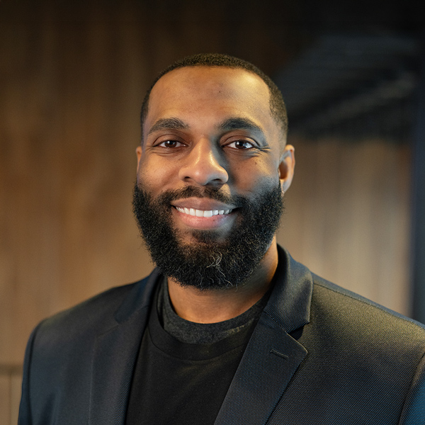 Kevin Banks, Talent Acquisition Manager at Arrow Senior Living, smiling in a black blazer against a softly lit wooden background.
