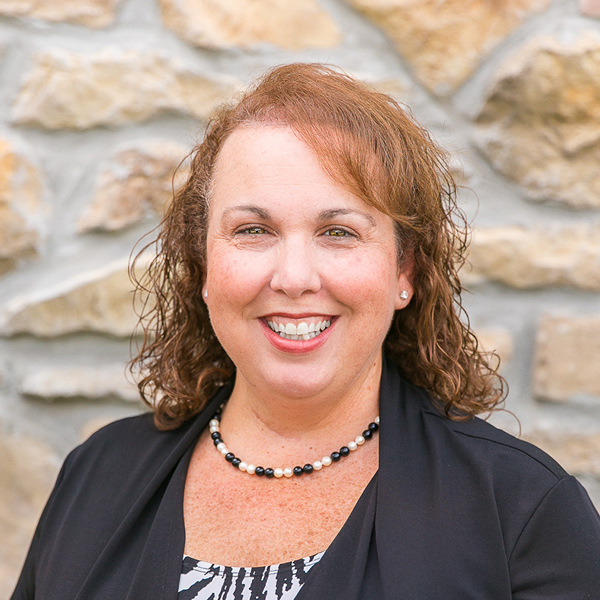 Terri Milsop, Regional Compliance Coordinator - Operations at Arrow Senior Living, smiling in front of a stone wall background, wearing a black jacket and beaded necklace.