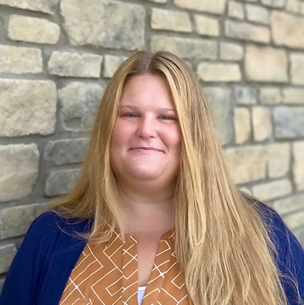 Kim Piaggio, Talent Acquisition Manager at Arrow Senior Living, smiling with long blonde hair, wearing a patterned top and blue sweater in front of a stone wall.