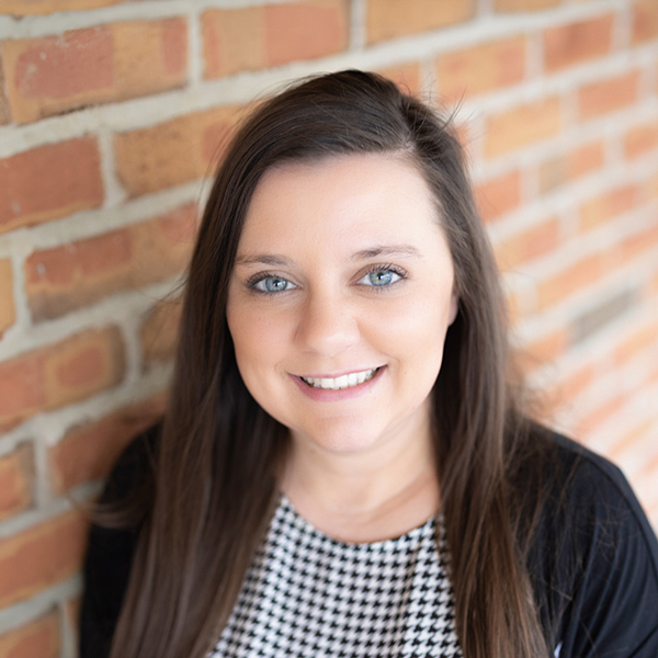 Sophie Rich, Engagement Specialist at Arrow Senior Living, smiles warmly while standing in front of a brick wall, wearing a patterned top and black cardigan.