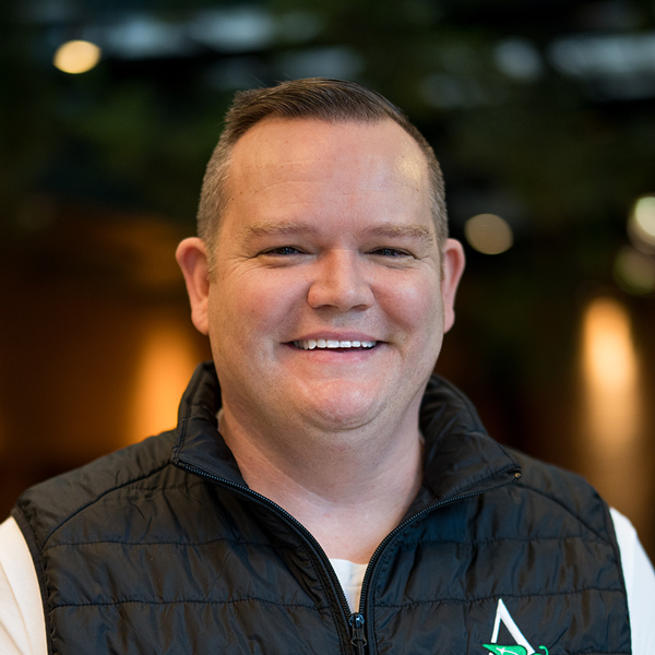 Tim Hart, Managing Director of Integration at Arrow Senior Living, smiles in a black Arrow-branded vest with a soft-focus background of warm interior lighting.