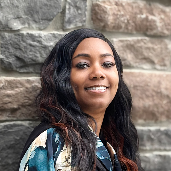 Che'Rri Colbert, Arrow Senior Living Regional Business Office Director, smiling confidently in front of a stone wall while wearing a patterned blouse with long, wavy hair.