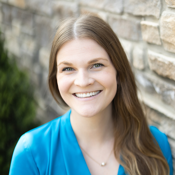 Annie Hoover, Engagement Specialist for Arrow Senior Living in the Ohio area, smiling warmly in a vibrant blue blouse with long light brown hair, standing next to a stone wall with greenery in the background.