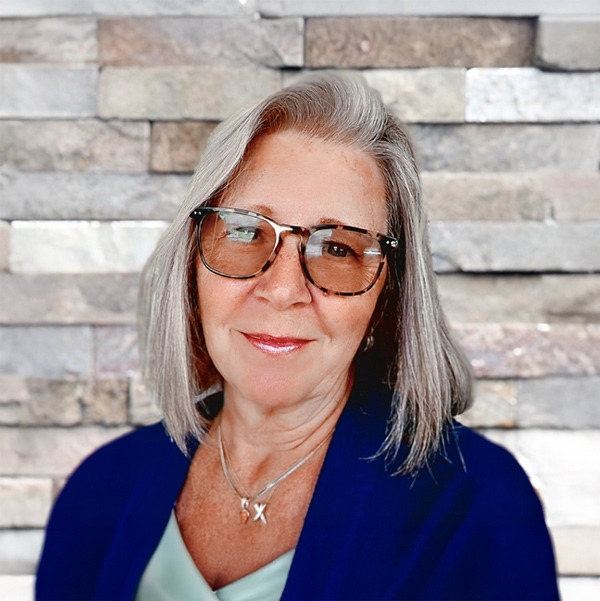 Debie Damas, Project Manager of the Wellness Team at Arrow Senior Living, smiling in a blue top and glasses, standing in front of a stacked stone wall.