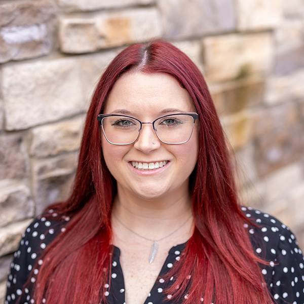 Kaitlynn Holley, Regional Memory Care Coordinator at Arrow Senior Living, smiling in front of a stone wall, wearing glasses, a black polka dot top, and a necklace.