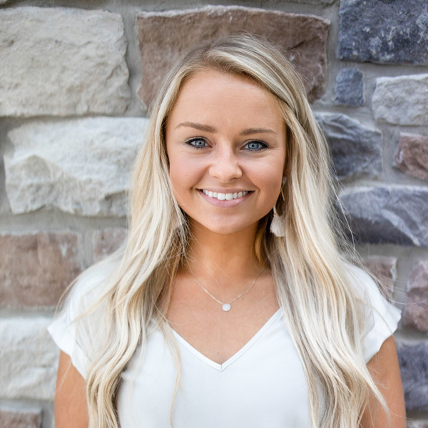 Laura Hogya, Engagement Specialist for Arrow Senior Living in the Ohio area, smiling in a white blouse with long blonde hair, standing in front of a multicolored stone wall.