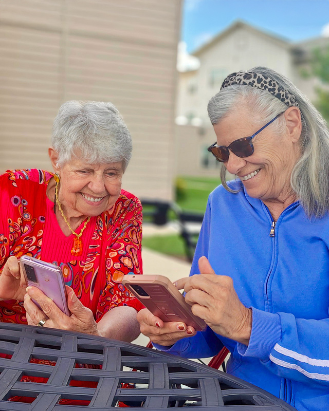 Two senior women sitting outside at a patio table, smiling and interacting with their smartphones on a sunny day.