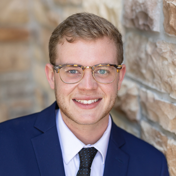 Tayte Moore, Arrow Senior Living Director for the Springfield, Missouri market, smiling in a navy suit, white shirt, and black polka dot tie, standing in front of a stone wall.