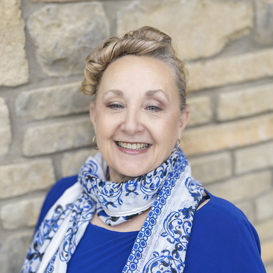 Erin Winstead, Arrow Executive Director in the Kansas City area for Arrow Senior Living, smiling warmly in a royal blue top and patterned scarf, standing in front of a tan stone wall.
