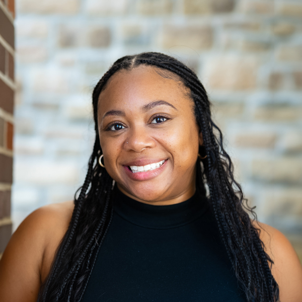 Ashley Moore, Regional Operations Specialist at Arrow Senior Living, wearing a sleeveless black turtleneck top with long braided hair, smiling in front of a blurred stone wall background.