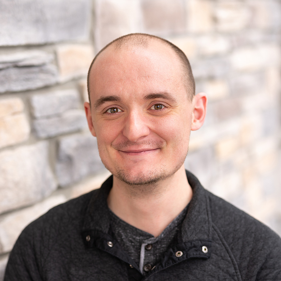 Austin Marsh, Resident Experience Coordinator at Arrow Senior Living, smiling in front of a neutral stone wall while wearing a textured dark pullover.