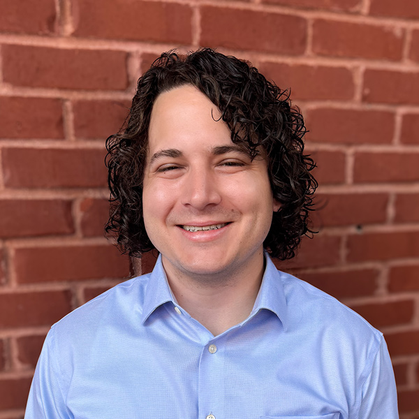 Cal Hockemeyer, Analytics Web Developer at Arrow Senior Living, wearing a light blue button-up shirt, standing in front of a red brick wall background.