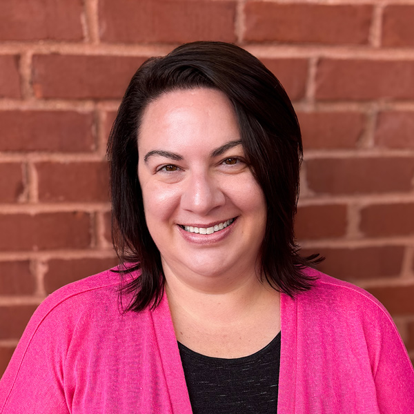 Kate Richardson, Financial Coordinator at Arrow Senior Living, wearing a bright pink cardigan over a black top, standing in front of a red brick wall background.