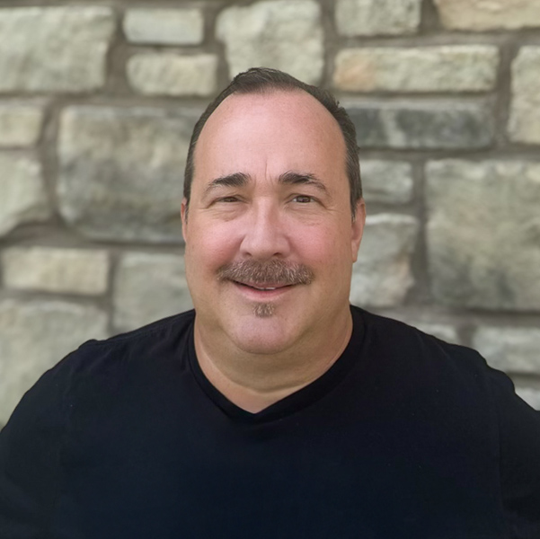 Michael Seelbach, Arrow Wellness Director for the Kansas City Region at Arrow Senior Living, wearing a black shirt and smiling in front of a stone wall background.