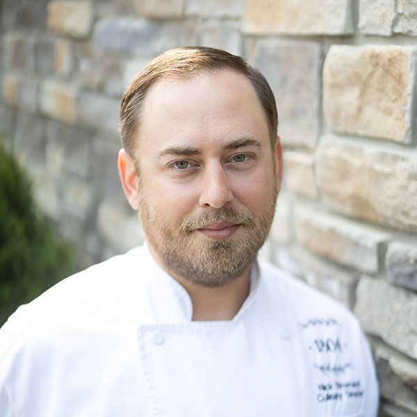 Nick Stromack, Regional Culinary Coordinator for the eastern market at Arrow Senior Living, smiling in chef’s coat against stone wall background.