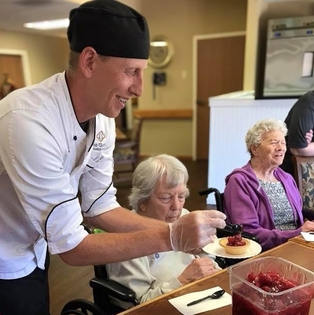 Chef in white uniform smiling while plating dessert for senior residents, serving fresh berries on cake during a culinary event in the senior living community.