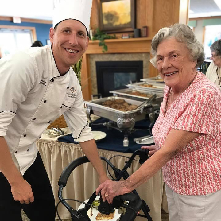 Chef in tall hat posing with a smiling senior woman at a buffet table, showcasing friendly dining experiences at the senior living community.