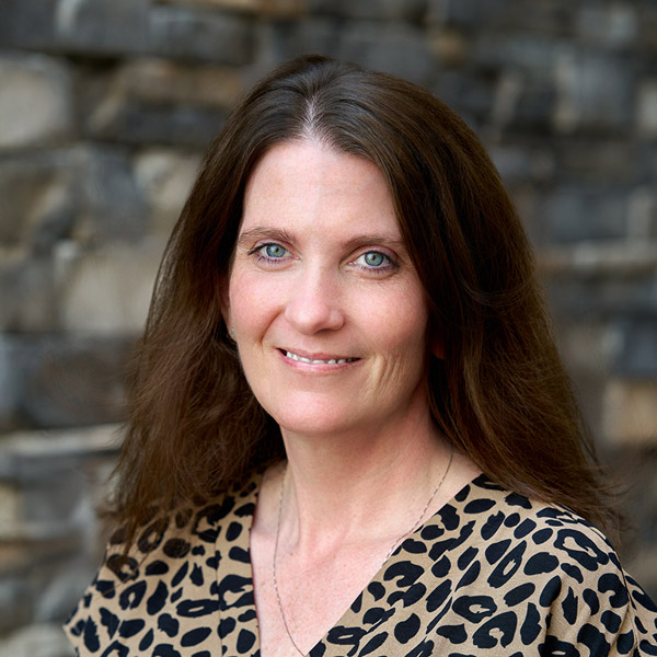 Holly Belanger, Senior Regional Wellness Coordinator at Arrow Senior Living, smiling in a professional headshot. She has long brown hair, blue eyes, and wears a leopard print blouse with a delicate necklace, standing in front of a stone wall background.