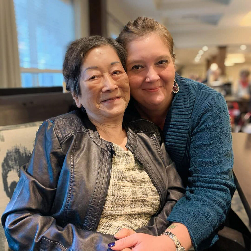 A smiling resident sits closely with a supportive team member, both sharing a warm embrace and joyful moment together inside the welcoming senior living community.