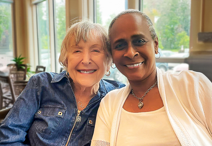 Two smiling women sit closely together by the window, enjoying each other’s company and a bright, welcoming moment in the community.