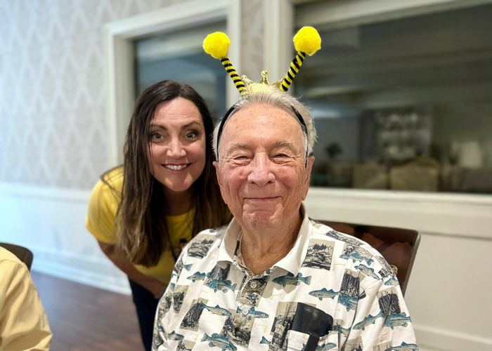 A cheerful senior man wears playful bee antennae while sitting beside a smiling team member during a themed event at the community.