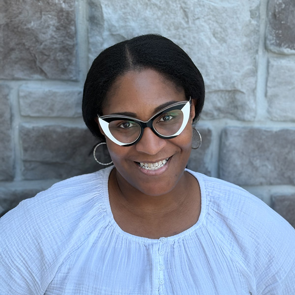 Brashawn Walker, Arrow Care Coordinator at Arrow Senior Living, smiling in a professional headshot. She wears bold black and white cat-eye glasses, hoop earrings, and a light textured blouse, standing in front of a gray stone wall background.